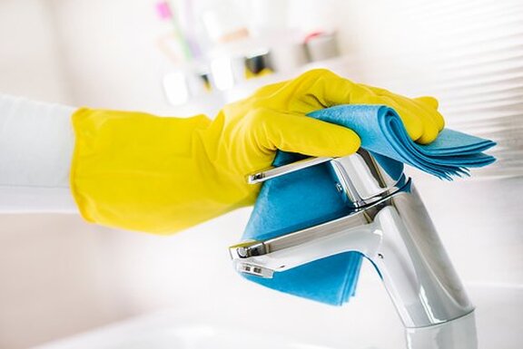 A close up picture of cleaning a bathroom tap. A hand in a yellow rubber cleaning glove is polishing a chrome bathroom sink faucet with a blue cleaning cloth.