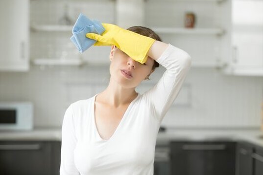 Frustrated woman in white tee shirt and yellow cleaning gloves holding a blue cleaning rag wiping her forehead with the back of her hand.