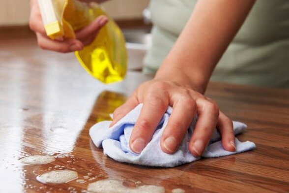 Close up image of cleaning a table top. One hand holds a blue cleaning cloth wiping soap suds off a table top while the other hand holds a spray bottle of cleaning solution.