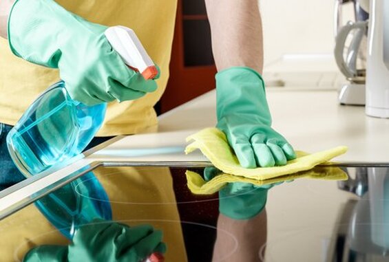 A closeup picture of hands in green rubber cleaning gloves deep cleaning a glass cooktop or stove in a kitchen. One hand holds a yellow cleaning cloth and is wiping the stovetop while the other hand holds a spray bottle of cleaning solution.