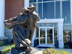 This is a picture of the main entrance to the West Jordan City Hall. There is a bronze statue of a man and woman in pioneer clothing in front of the entrance door, which has white lettering which reads, 